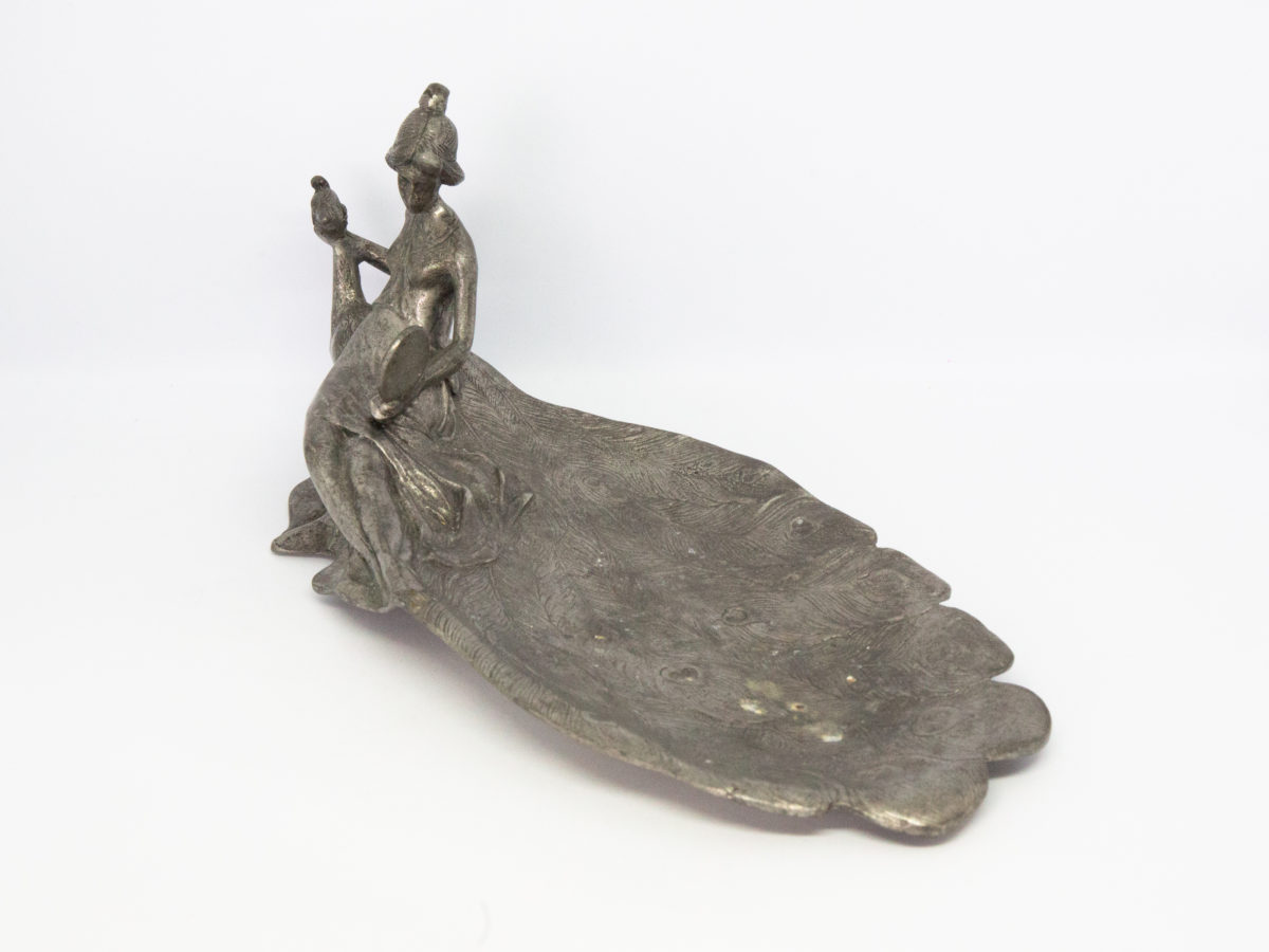 Antique Art Nouveau pewter pin tray. A rare and very lovely decorative pewter pin tray of a young lady seated on the back of a peacock gazing into a mirror. The tray area is plumes of the peacocks fanned out feathers. Lots of attention to detail in this piece. Is she looking at herself or the peacock in the mirror? 95% pewter (Zinn 95%) Photo of pin tray at an angle so the tail of peacock is in the foreground and to the bottom right of photo and girl and head of peacock is in the top left of photo.