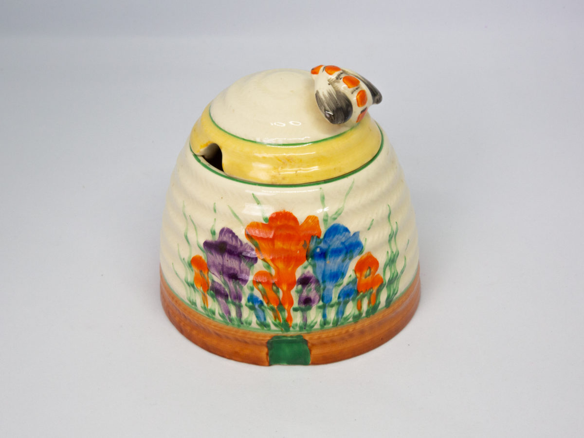 Clarice Cliff 'Autumn Crocus' honey pot. A sweet and classic Clarice Cliff design honey pot hand-painted in the Autumn Crocus pattern. Shaped in the form of a beehive with a bee handle to the lid. In excellent condition throughout with only minor stain on small inside area. Signed Clarice Cliff to the base. c Late 1930s. Base of pot measures 96mm in diameter. Height excluding bee handle 85mm. Main photo of pot with lid in place, bee handle to the right, spoon hole on lid to the left and green painted hive entrance at the bottom.
