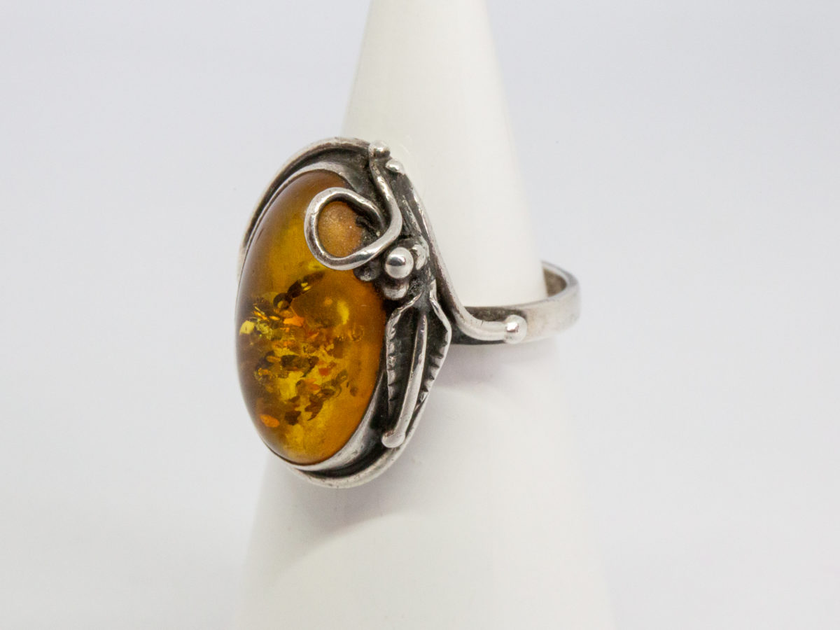 1960s vintage silver and amber ring. Pretty honey amber set on sterling silver with an Art Nouveau style decorative touch to one side. Worn hallmark to the outside band of ring at the back. Ring front measures 25mm by 20mm. Ring size O.5 / 7.5. Photo of ring displayed on a cone shaped stand and seen with ring front facing left of photo.