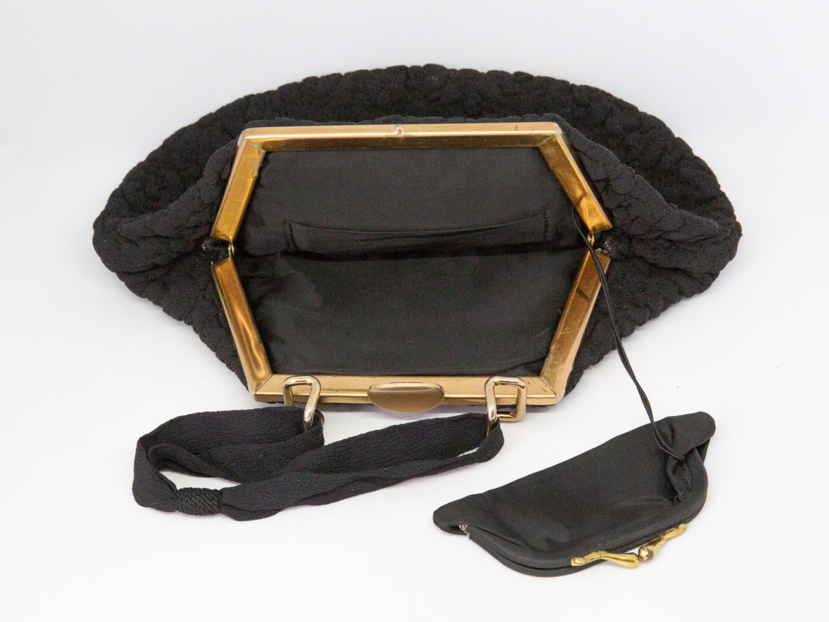 40s Black fabric bag with brass clasp. A lovely 40s bag in black fabric with a floral accent. Interior has a small pocket and a tiny coin purse attached to string. Brass clasp and short black fabric handle completes the look. Drop length from handle to bottom of bag is 300mm. Photo of bag seen from the frame end with the clasp open and small attached coin purse laid outside of the handbag.