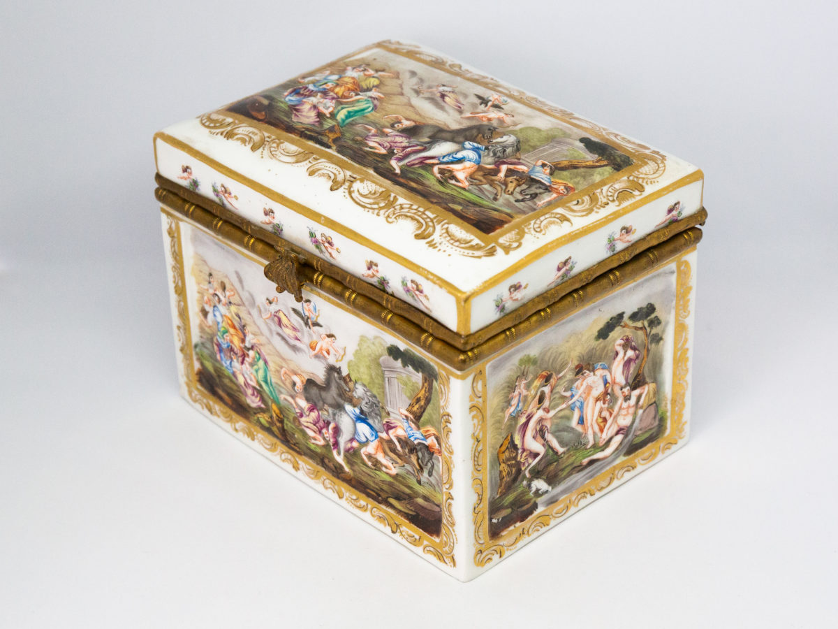 Late 19th century Naples porcelain box. A heavy porcelain box moulded in high relief with scenes of scantily clad men and women disporting themselves in various ways. Beautifully hand-painted in bright vibrant colours and finished off with gilt accent. Photo of box set diagonally with clasp front facing left and showing the side, the front and the lid decorations.