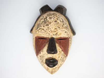 Small vintage wooden African mask. Lovely African mask carved from one piece of wood and coloured in natural pigment. Has a sweet smiling facial expression-even looks as though it is blowing kisses from certain angles- and an unmistakeable aroma of bonfires. Main photo of mask laid on a flat surface and looking straight down from above.