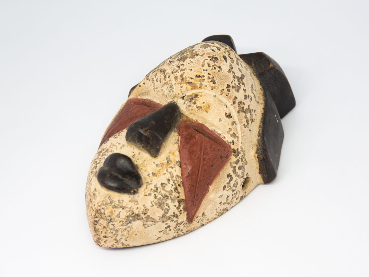 Small vintage wooden African mask. Lovely African mask carved from one piece of wood and coloured in natural pigment. Has a sweet smiling facial expression-even looks as though it is blowing kisses from certain angles- and an unmistakeable aroma of bonfires. Photo of mask on a flat surface and laid diagonally with chin in the bottom left corner and top of head to the top right of photo