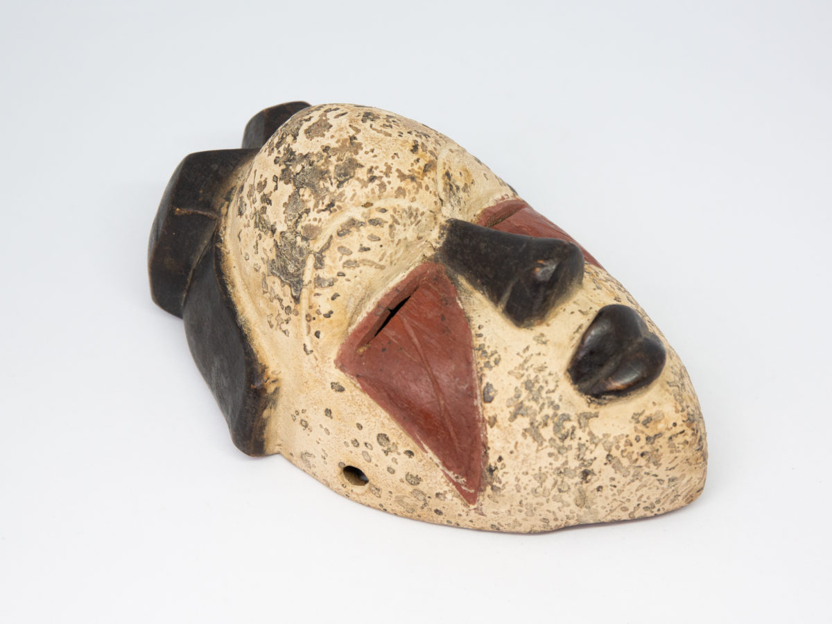 Small vintage wooden African mask. Lovely African mask carved from one piece of wood and coloured in natural pigment. Has a sweet smiling facial expression-even looks as though it is blowing kisses from certain angles- and an unmistakeable aroma of bonfires. Photo of mask on a flat surface and laid diagonally with chin in bottom right of photo and top of head in top left,