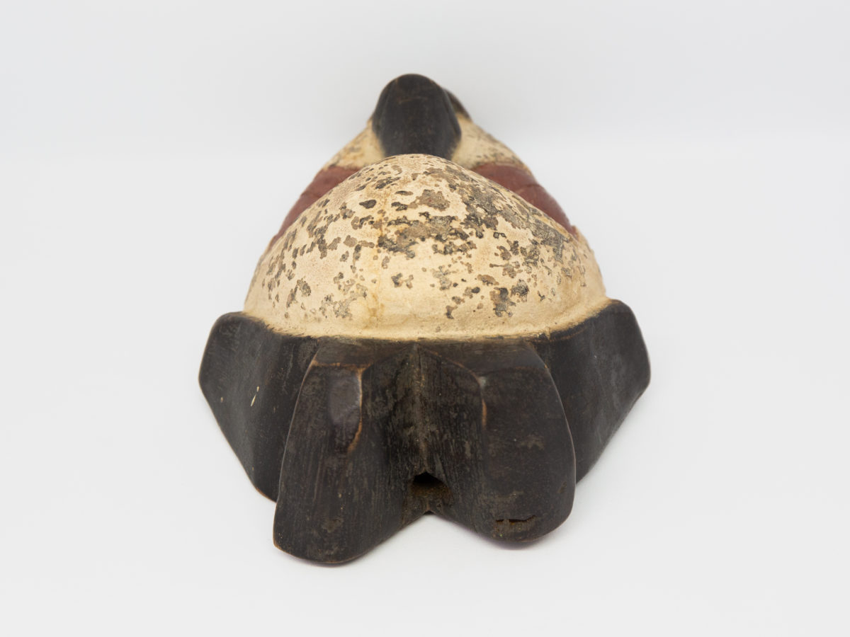 Small vintage wooden African mask. Lovely African mask carved from one piece of wood and coloured in natural pigment. Has a sweet smiling facial expression-even looks as though it is blowing kisses from certain angles- and an unmistakeable aroma of bonfires. Photo of mask on a flat surface and seen with top of head in foreground.