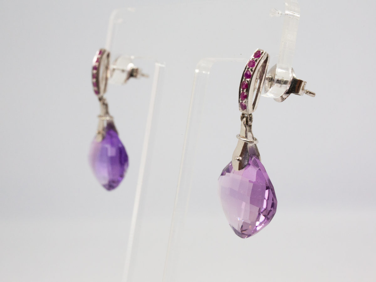 Modern sterling silver earrings with amethyst and rubies. Pretty pair of modern sterling silver dangle earrings with generous size cushion cut amethyst stones and 5 small rubies to each. Hallmarked 925 for sterling silver to the back and on butterflies. Box included. Amethyst measures 15mm square and earring dangle length is approximately 35mm. Earrings weigh 6.1gms. Photo of earrings displayed on a Perspex stand and seen from a slight side angle with earring fronts facing left of photo.