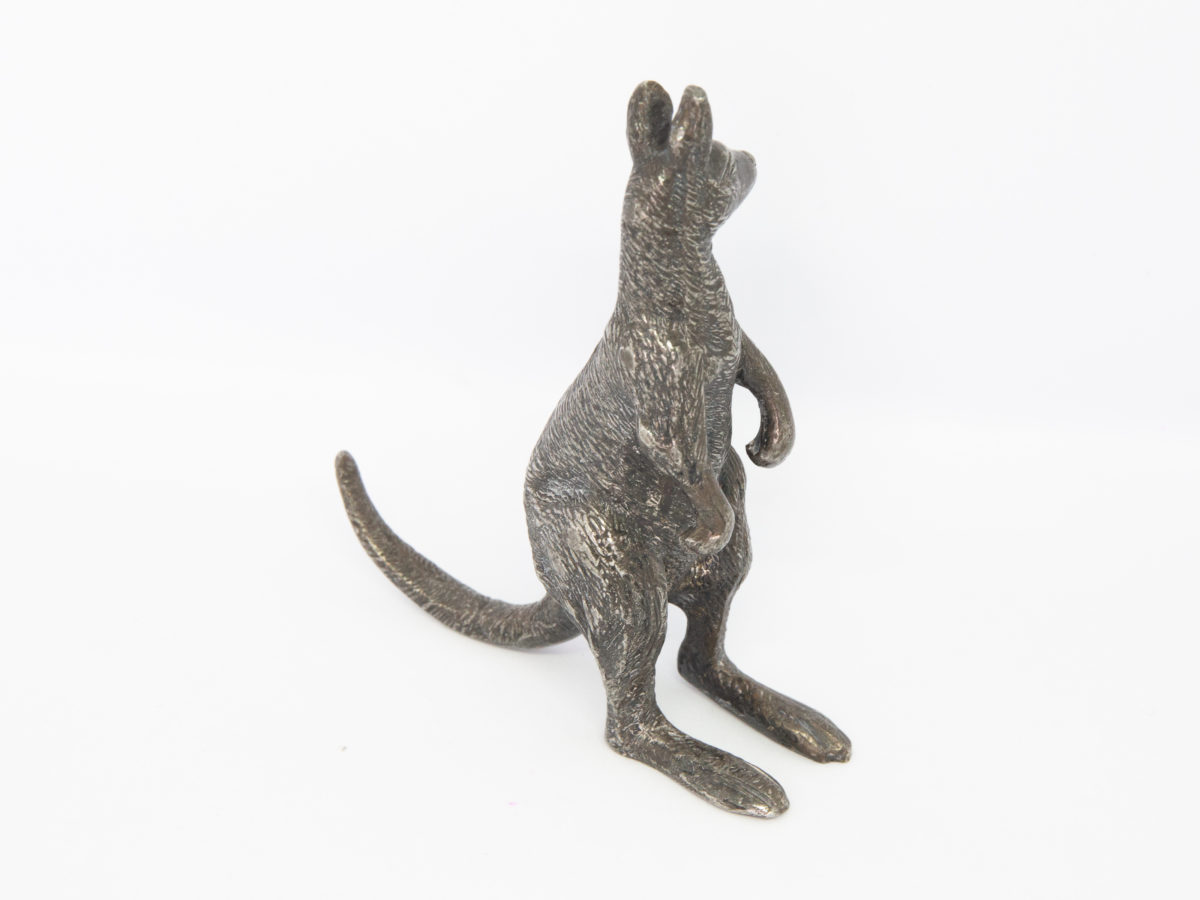 Antique Stewart Dawson kangaroo ornament. A small heavy kangaroo in standing position by Stewart Dawson. Assumed solid silver plate although no visible hallmarks whatsoever. Signed Stewart Dawson to the base of tail. c1900s Measures 85mm at longest from tip of foot to tail, 28mm at widest across hips and 98mm at tallest from ground to tip of ear. Photo of kangaroo posed with tail pointing top left of photo and kangaroo facing top right.