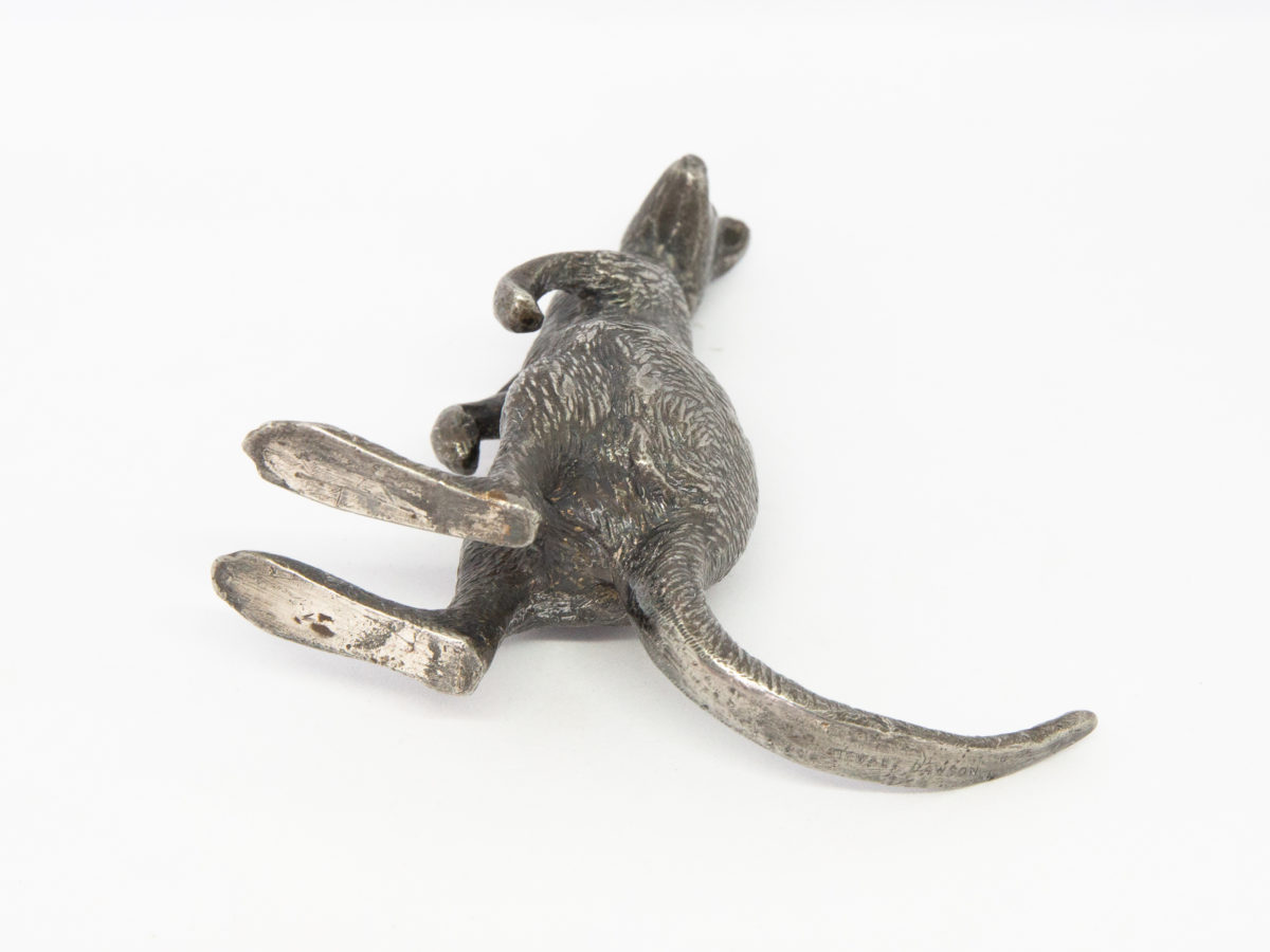 Antique Stewart Dawson kangaroo ornament. A small heavy kangaroo in standing position by Stewart Dawson. Assumed solid silver plate although no visible hallmarks whatsoever. Signed Stewart Dawson to the base of tail. c1900s Measures 85mm at longest from tip of foot to tail, 28mm at widest across hips and 98mm at tallest from ground to tip of ear. Photo of kangaroo laid on its side showing the bottom of the tail and feet.