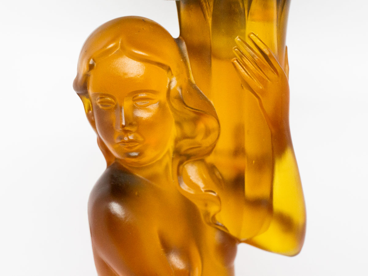 Art Deco glass lamp and shade. Stunning amber glass lamp of semi naked seated lady holding a multi dimensional amber glass shade. The shade is a slightly lighter colour to the main lamp. Lamp base measures 145mm in diameter. Close up photo of face of lady.