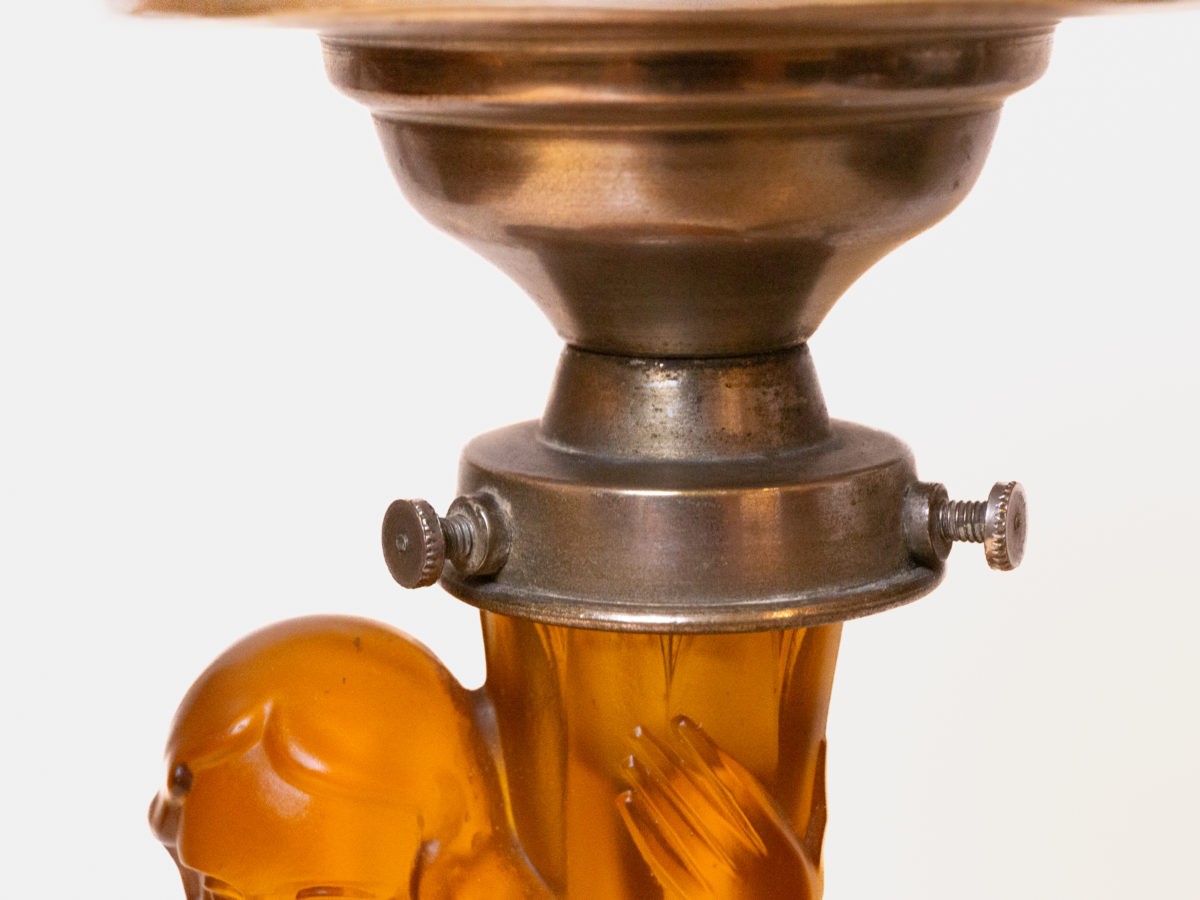 Art Deco glass lamp and shade. Stunning amber glass lamp of semi naked seated lady holding a multi dimensional amber glass shade. The shade is a slightly lighter colour to the main lamp. Lamp base measures 145mm in diameter. Close up photo of the area connecting lady to the lamp shade area.