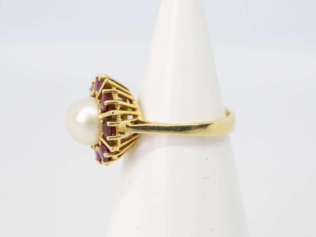Vintage 14 karat gold ring with pearl and rubies. Pretty ring with a central pearl framed by 8 round cut rubies in a star/sun burst design on an open prong cathedral setting. Ring size N.5 / 7. Ring weight 5.6gms. Photo of ring on a cone shaped display stand with ring front facing left and showing the open prong cathedral setting.