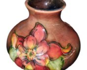 Moorcroft vase in flambe orchid pattern. Beautiful little vase, highly decorative in a rust flambe colour with orchids in green and yellow decorating the body. Main photo of vase seen from a near eye level angle showing the orchid decoration on the side.