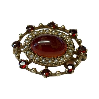 Vintage 9 karat gold oval brooch set with a garnet cabochon to the centre. A frame of seed pearls surround the central garnet then an outer ring of gold and round cut garnets border the whole brooch. Hallmark worn but visible for London assay. Brooch measures 30mm by 26mm. Box included (may not be box shown) Main photo of brooch front with garnet cabochon to centre, seed pearls framing it and an outer frame of small garnets on the outside border.