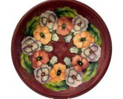 The Moorcroft Pansy plate is a beautiful example of the work of William Moorcroft. The plate has a vibrant red background and a series of colourful pansies decorating the inside. Main photo of plate seen upright and from the front.