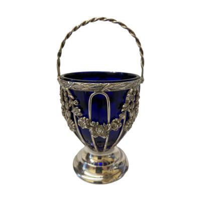 Antique Chester silver basket with glass lining. Beautiful small basket with sterling silver floral garlands to the side and a twist handle. The sterling silver basket is lined with Bristol blue glass.  Fully hallmarked to the rim for Chester assay c1908 with short hallmark to one handle end. Made by Haseler Brothers. Measures 58mm in diameter at base and 70mm at top. Height is 135mm with handle fully extended and 90mm to top of basket. Main photo showing basket with handle raised like a rainbow and a floral garland like a reflection of the handle below