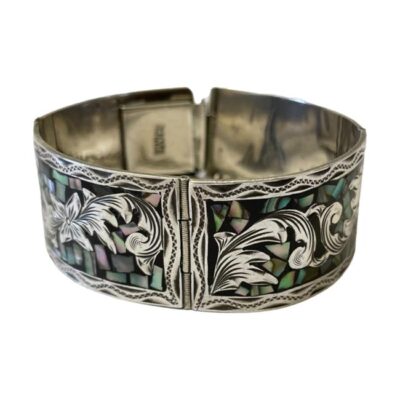 Mexican panel bracelet inlaid with abalone. A vintage Mexican sterling silver panel bracelet with a floral theme and finished with small pieces of inlaid abalone pieces to enhance & compliment the intricate silver work. Hallmarked sterling Mexico and 925. Wide safety chain. One panel appears slightly misshapen but is not noticeable and does not affect clasp. With clasp undone & safety chain extended, the width of opening is maximum 94mm. Main photo of bracelet seen from a slightly raised eye level focusing on the 2 panels on view at front. The clasp area and the hallmark is visible in the background.